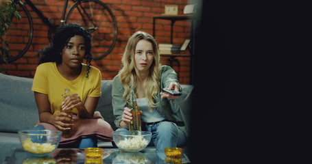 Two mixed-races pretty girls sitting on the couch in the living room and changing channels with remote control while choosing what to watch on TV.