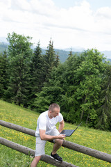 man sits on a wooden fence and works with a laptop near the field and coniferous forest. Vertical frame