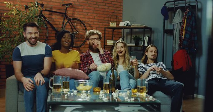 Multiethnical young and cheerful men amd women watching comedy movie on TV together in the loft styled living room with beer and snacks and laughing laud.
