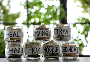 Glass jars with money for different needs on table against blurred background