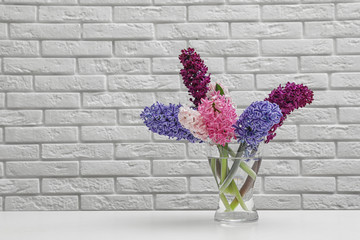 Beautiful hyacinths in glass vase on table against brick wall, space for text. Spring flowers