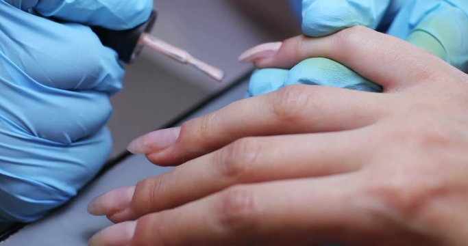 Close-up view of nail master in blue gloves covering nails with colorless varnish. Young woman in nail salon.