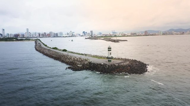 Drone view distancing of a lighthouse of Farol Do Molhe Da Barra De Itajaí, Santa Catarina, Brazil, showing the city on the horizon and waves craching on the concrete blocks.