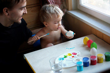 Fototapeta premium Father and his toddler blond son dyeing Easter eggs together at home, they laugh and have fun together