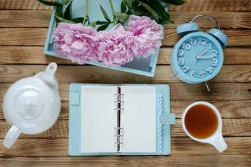 Spring tea.Spring season Flat lay.blue notebook blue alarm clock, bouquet of pink peonies, white teapot and cup of tea on wooden board background.Spring time