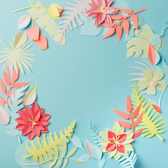 Fototapeta na wymiar Colourful handmade tropical paper flowers and leaves on blue pastel background with copyspace in the middle, summer spring flower concept, papercraft origami idea