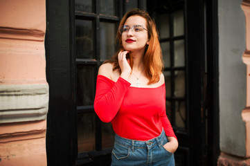 Attractive redhaired woman in eyeglasses, wear on red blouse and jeans skirt posing at street against old wooden door.