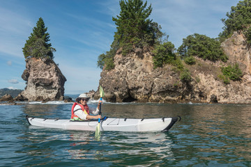 Adult, female kayaker paddling along the rocky coast of Coromandel, New Zealand, with cliffs, stacks and trees behind on the shore.