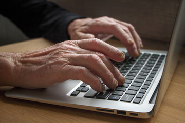 The hands of an elderly person working on a laptop keyboard, close up photo.