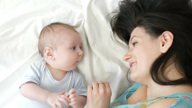 A beautiful young mother and a funny two-month newborn baby are lying on the bed and looking at each other. Mom gently strokes her baby.