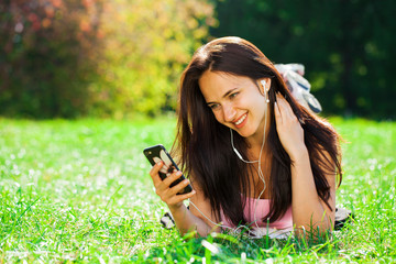 Young woman in dress lies on green grass and listens to music in headphones through smartphone.
