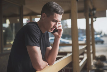 A handsome, slender young man in a black t-shirt on the street uses a smartphone and speaks on it