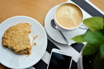 cup of coffee and cookie on wooden table with plant and white smartphone