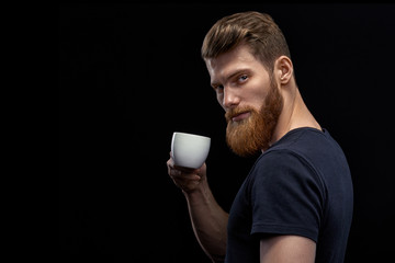 Beautiful young bearded man drinking coffee and looking at camera Resting handsome perfect hairstyle man drinking espresso coffee. Man holding cup of coffee in hand over black background.