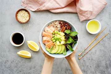 Girls' hands holding salmon poke bowl Salmon poke bowl with salad, avocado, edamame beans, pineapple, sesame and brown rice Traditional Hawaiian dish
