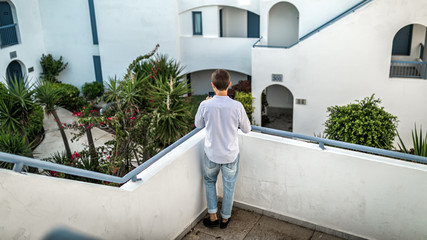 Young guy stands with his back on balcony, terrace overlooking garden with palm trees in Greece. White background, space for text
