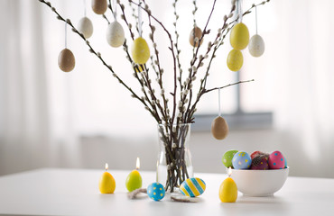 holidays and object concept - pussy willow branches decorated by easter eggs in vase and candles on table