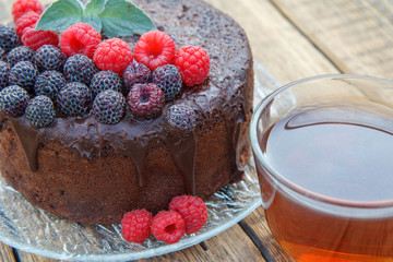 Homemade chocolate cake decorated with black and red raspberries on glass plate with cup of tea.