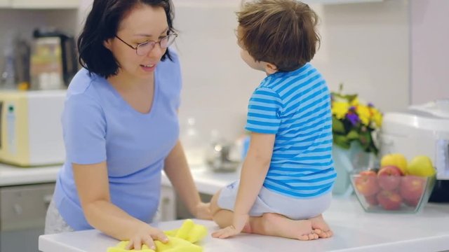 Beautiful smiling mixed race mother washing table. Sweet little baby son playing near and talking with mom. Young cheerful mom keeping house clean. Slow motion.