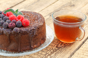 Homemade chocolate cake decorated with black and red raspberries on glass plate with cup of tea.