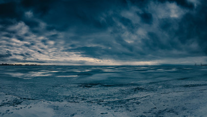 Naklejka na ściany i meble Frozen sea landscape with dark dramatic cloudy sky over water covered with snow and starting to melt. Seascape