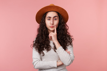 Close up of young lady with a hat on head, one finger touches cheek, standing isolated over pink background, looked up and thinking.