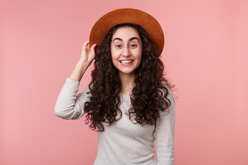 Image of happy young woman with curly hair, holds hat on head, wears casual white longsleeve, smiles broadly, isolated over pink background. Positive emotion concept.