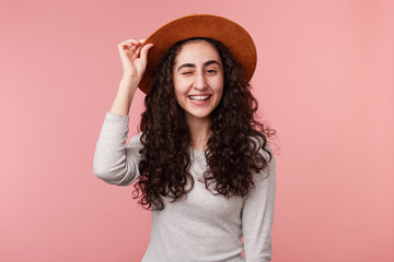 Photo of happy young woman with curly hair, holds hat on head and winking, wears casual white longsleeve, isolated over pink background. Positive emotion concept.