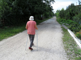 Nordic walking, therapy for health, retirement in the village. Old woman walking with sticks on rural street in summer, sports exercises for healthy spine and joints