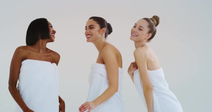 Slow motion of beautiful young smiling women of different ethnicities with perfect firm and slim body in white bathing towels whispering their beauty secrets in the ear and having fun.