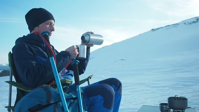 Camping in winter mountains. Climber mountaineer csitting in chair ooking outdoor in winter. Tourist backpacker travelling in winter, high snow mountains , trekking and camping equipment, camping tent