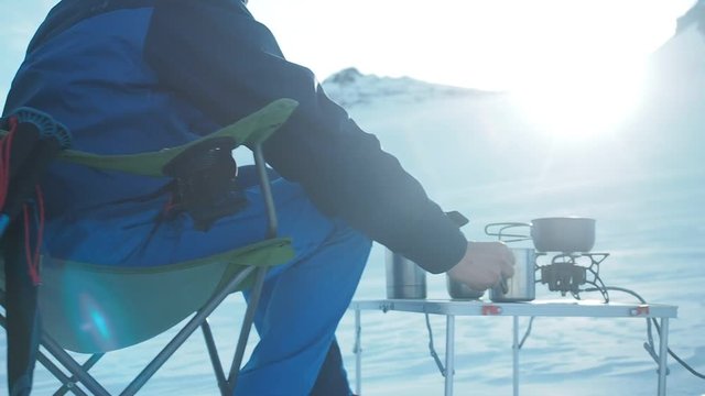 Camping in winter mountains. Climber mountaineer csitting in chair ooking outdoor in winter. Tourist backpacker travelling in winter, high snow mountains , trekking and camping equipment, camping tent