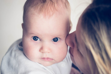 Mum kisses a baby baby in a cheek. The girl has a funny look. Caucasian child, blue eyes and red hair. Mom is not in focus.