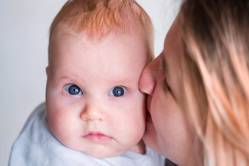 Mum kisses a baby baby in a cheek. The girl has a funny look. Caucasian child, blue eyes and red hair. Mom is not in focus.
