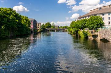 Fototapeta na wymiar River Corrib landscape in Galway, Ireland, as seen from the Wolfe Tone Bridge under a beautiful blue sky on a sunny summer day, bordered by houses and green trees.