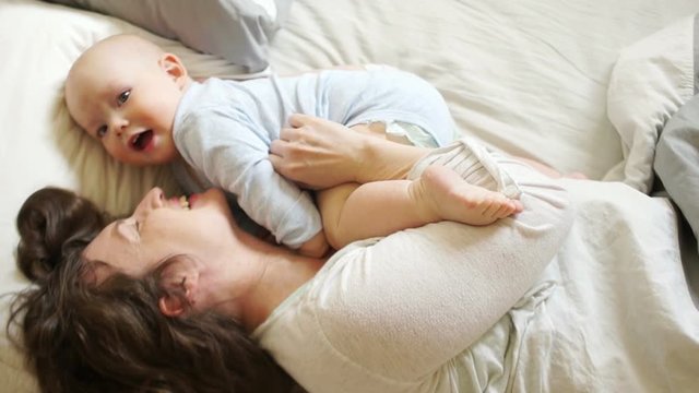 Woman tickling and hugging her year-old son. Sunny family morning mom and baby in bed. Mothers Day
