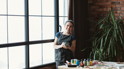 Break time. Rest and relaxation. Delight. Woman artist with mug smiling looking at camera. Art studio workspace.