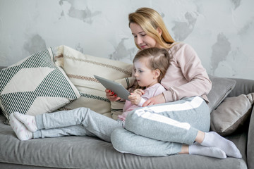 Beautiful young mother and her little daughter are watching movies together and playing on the tablet while sitting on the sofa. Mom hugs daughter. Maternal care and love. Horizontal photo