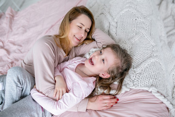 Obraz na płótnie Canvas Beautiful young mother and her little daughter are lying together on the bed in the bedroom, laughing, hugging and having fun. Maternal care and love. Horizontal photo