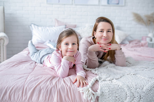 Beautiful young mother and her little daughter are lying together on the bed in the bedroom, playing, hugging and having fun. Maternal care and love. Horizontal photo