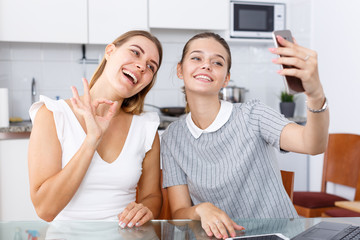 Two young girlfriends making selfie by phone and smiling at table