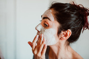 Young woman applying a revitalizing white mask on her face at home. Beauty treatment, skin care,...