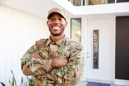 Soldier in camouflage standing outside modern house with arms crossed smiling to camera, close up