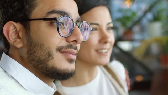 Close-up rack focused shot of young asian woman and middle eastern man in eyeglasses smiling, talking and embracing while looking through the window in cafe