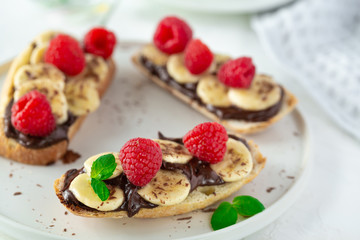 Front view of toast bread with topping - chocolate paste, bananas, raspberries and mint on white background with flowers and teapot. Tasty sweet summer breakfast with toasts and cup of tea