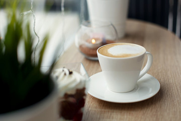 White cup of hot cappuccino on white saucer and red velvet dessert on wooden bar table next to window. Cold snowy weather outside