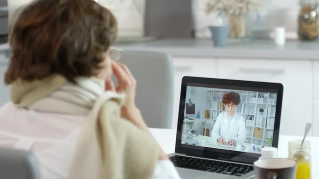 Medium shot of Caucasian woman sitting at laptop in kitchen and telling online female doctor about her sickness symptoms