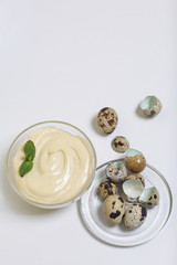 Handmade mayonnaise using quail eggs. In a glass container, decorated with greenery. Near quail eggs and shell from them. On a white background.