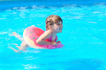 Cute smiling little girl with inflatable donut circle learns to swim in swimming pool on hot sunny day. Healthy and happy childhood concept.