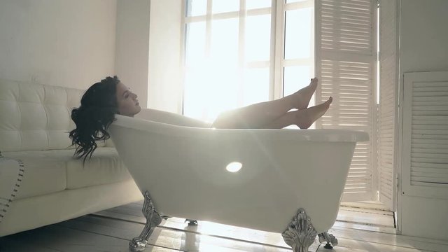 beautiful dreaming brunette girl with bare feet lies in stylish retro bath in modern living room on day